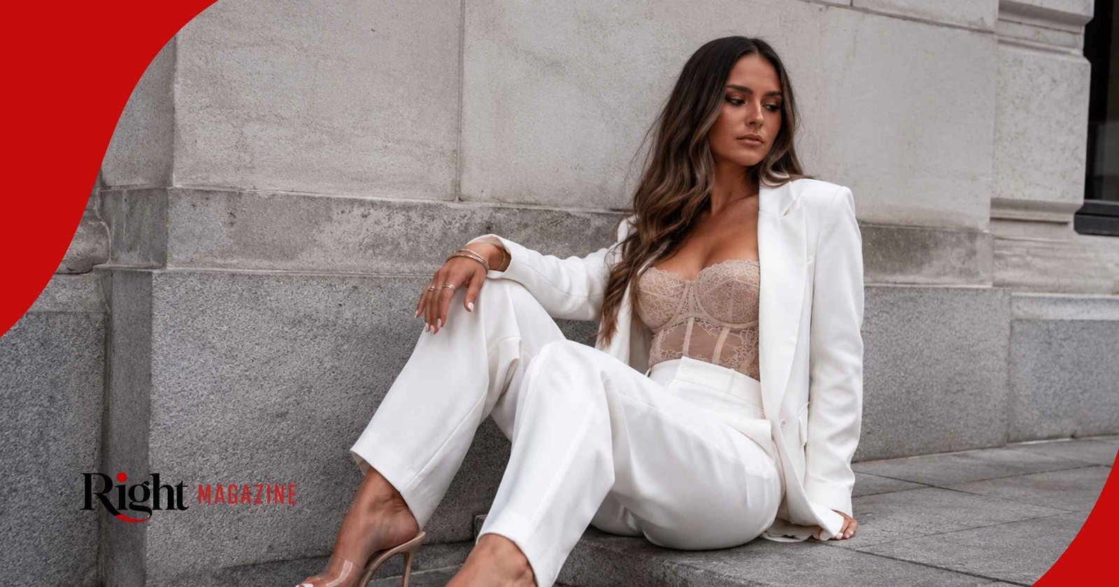 Leah Taylor sitting on a stone step, wearing a chic white pantsuit over a sheer lace bodice, looking away thoughtfully.