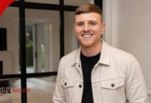 Tommy Mallet smiling while wearing a light cream denim jacket over a black t-shirt inside a modern house.