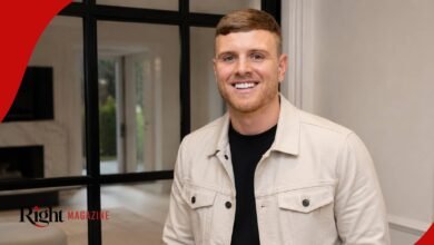 Tommy Mallet smiling while wearing a light cream denim jacket over a black t-shirt inside a modern house.