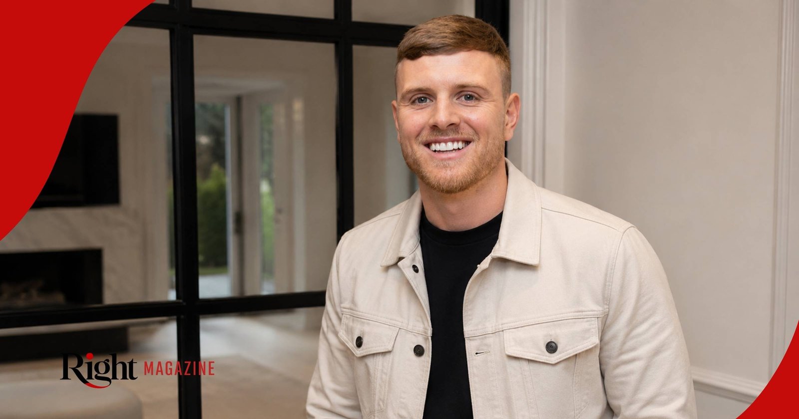 Tommy Mallet smiling while wearing a light cream denim jacket over a black t-shirt inside a modern house.